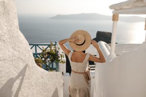 Lady holds straw hat. Woman in beige dress with bag goes down to sea. Girl in stylish outfit walks between white walls.
